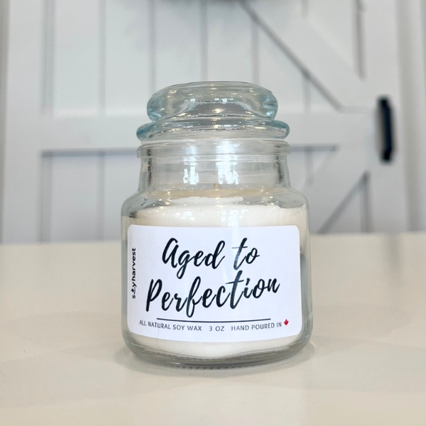 Glass jar candle labeled 'Aged to Perfection' on a white surface with a neutral background.