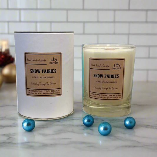 A beige candle with a label reading 'Snow Fairies' placed inside a white cylindrical container, which is part of a drinking glass set. The set is displayed on a wooden platform.